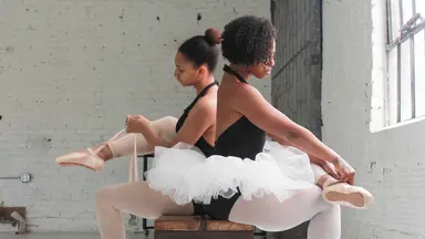 Two woman in tutus sitting on a bench and lacing up ballet shoes