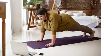 Woman doing a livestream yoga class on a mat in her home next to her computer